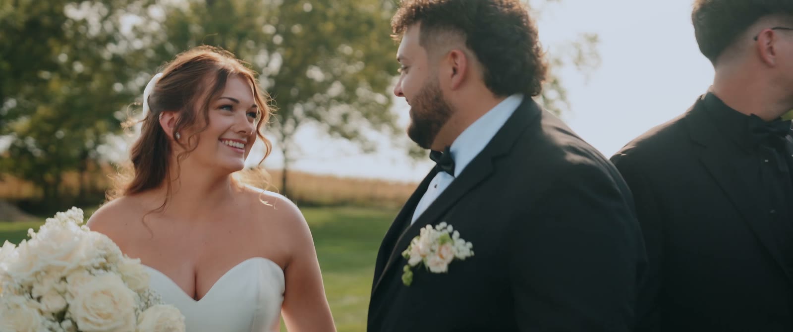Jacey and Ryan smile at each other outdoors as she holds a white rose bouquet.