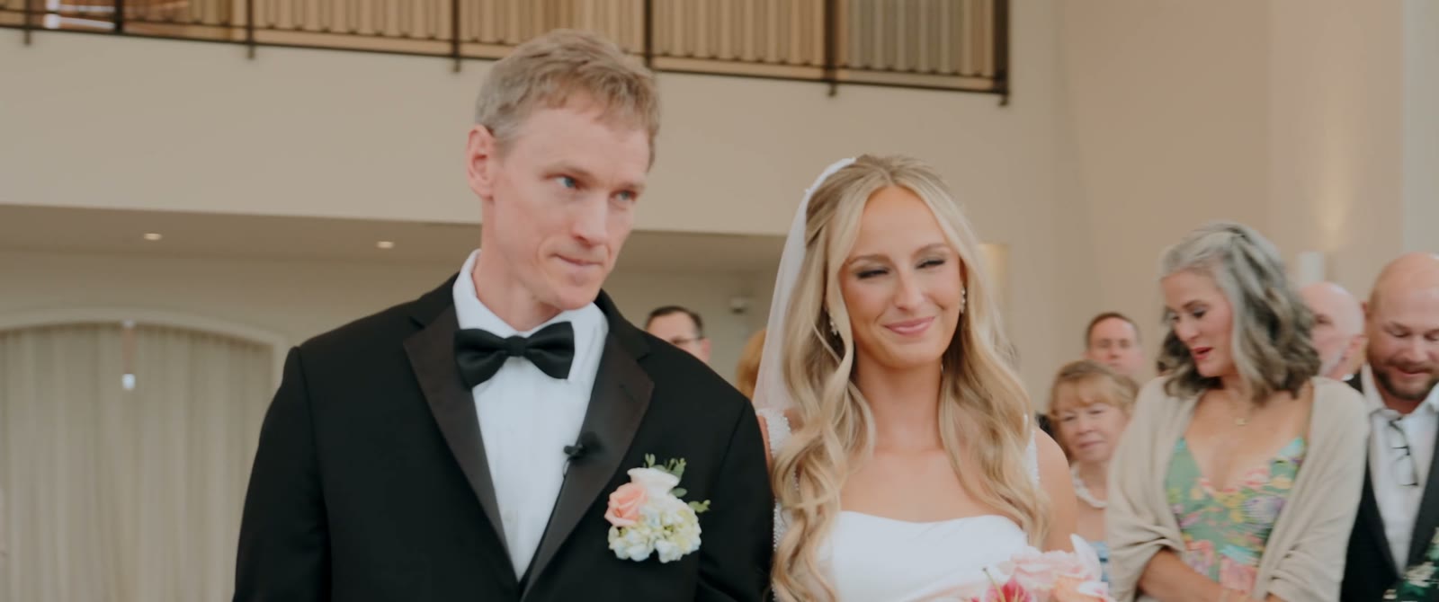 The bride smiles with eyes closed as she walks the aisle beside a man in a black tuxedo.