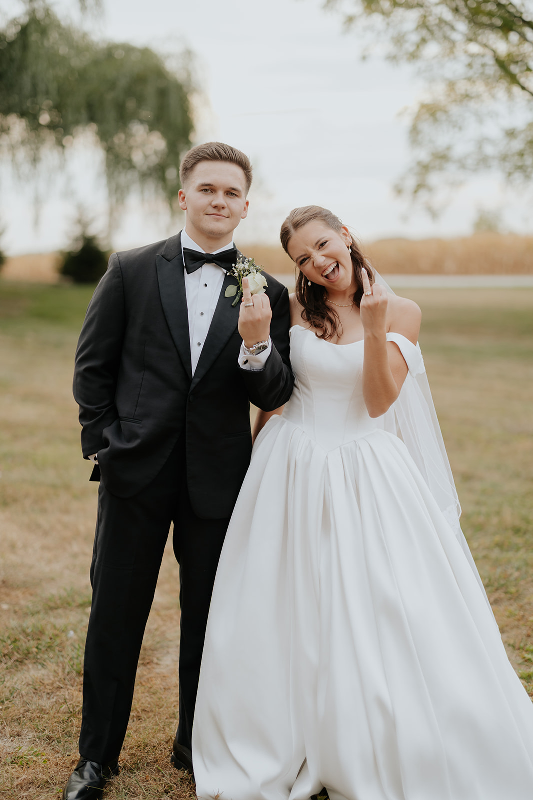 Regan and Reyn pose in a field, the bride laughing as both playfully show their rings.