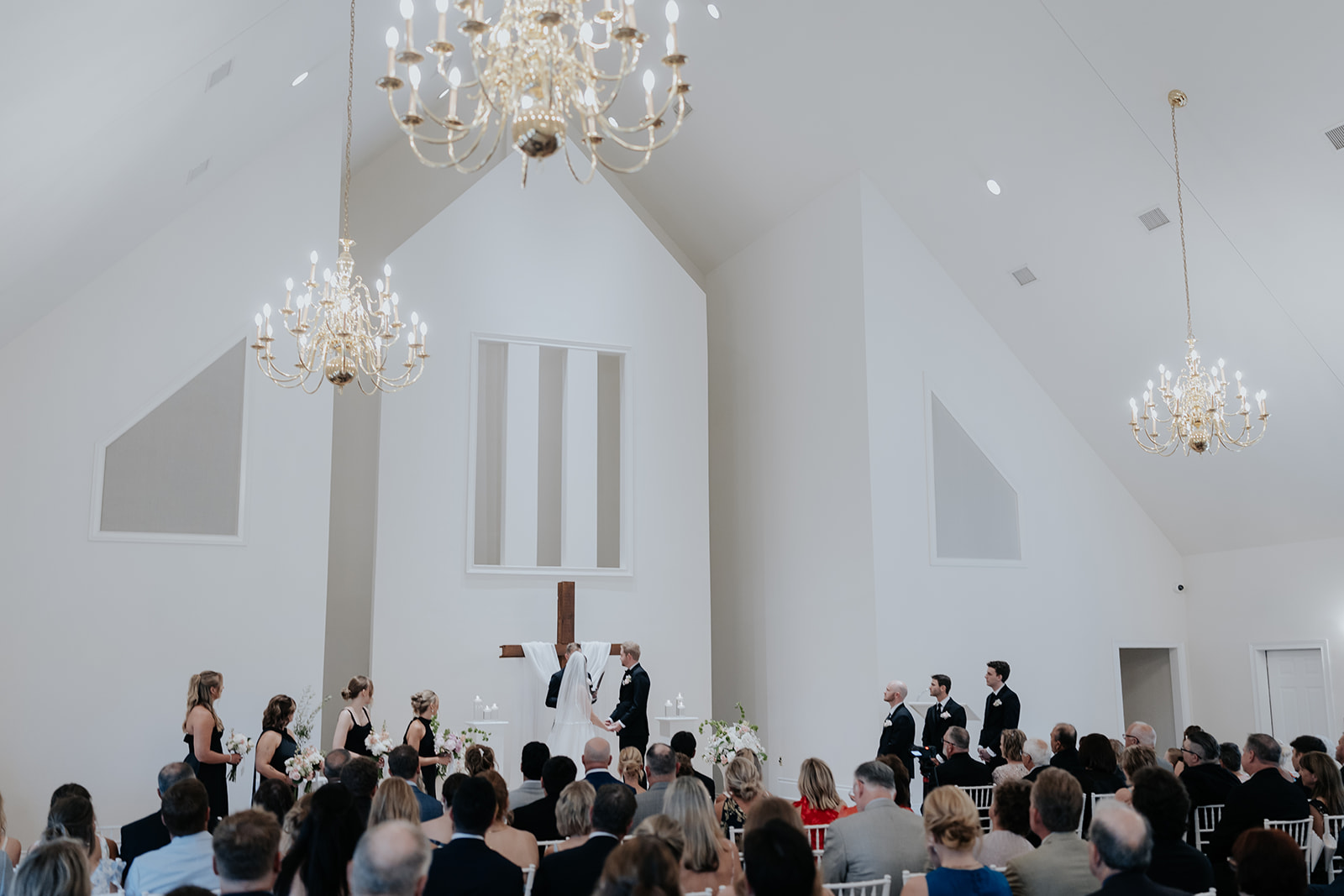 Wide view of a ceremony in a white chapel with chandeliers, the wedding party and guests watching.