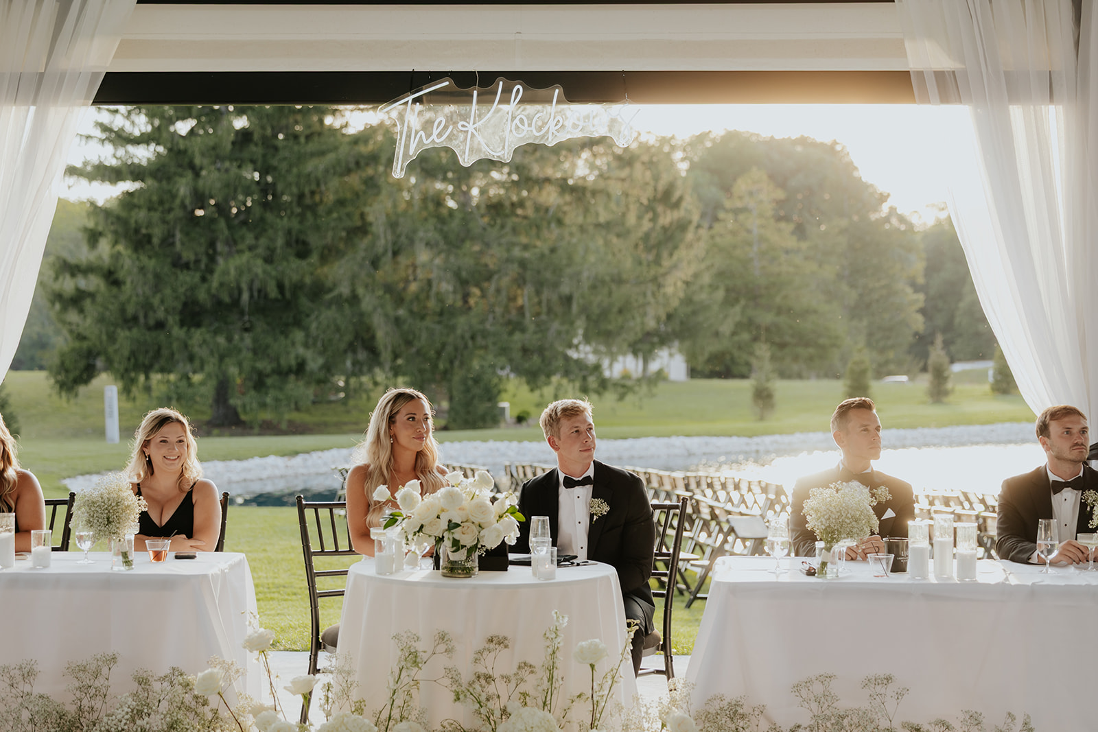 The couple and wedding party sit at reception tables under a neon sign with a pond view behind.