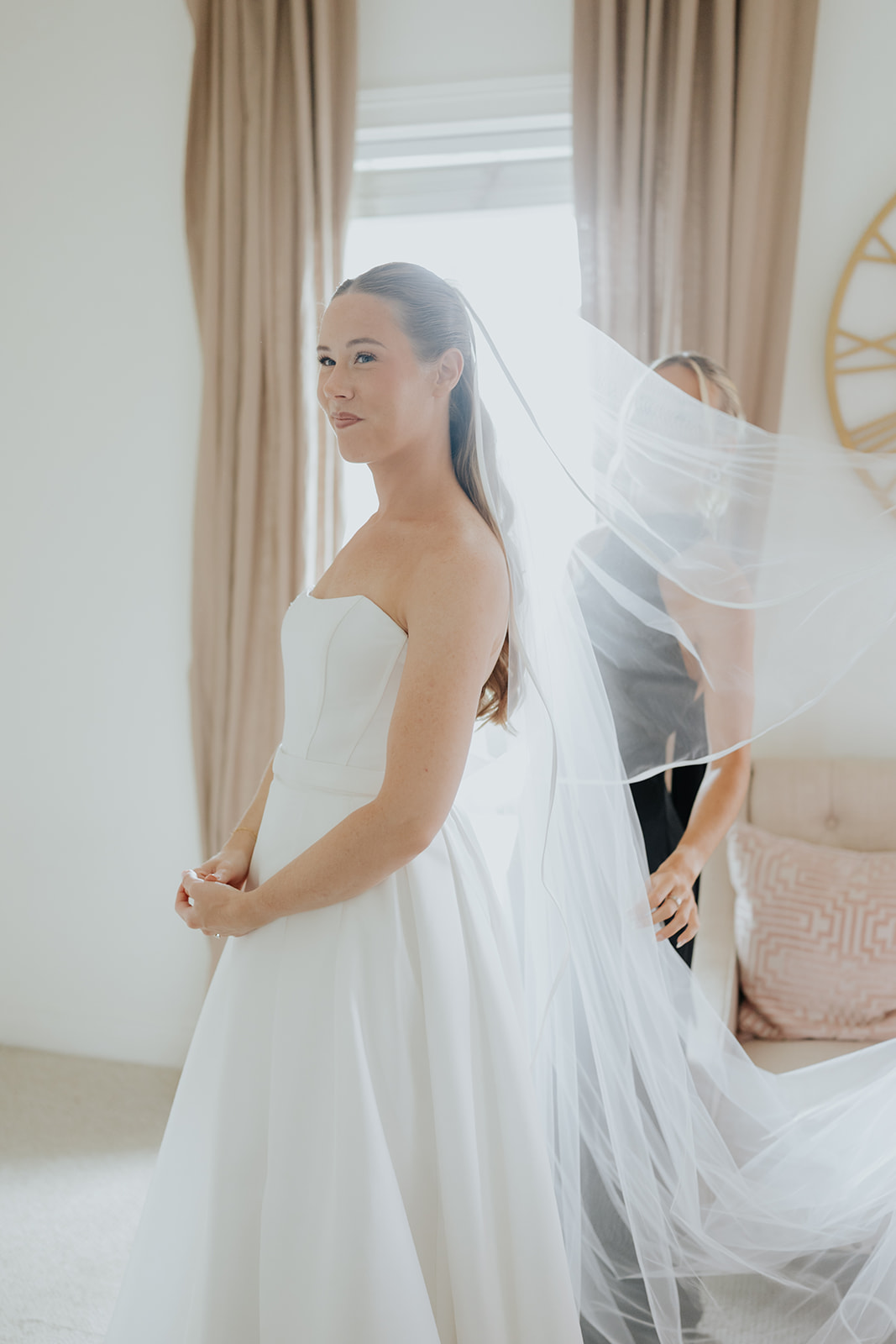 Brooklyn stands by a sunlit window as her long veil is arranged behind her while getting ready