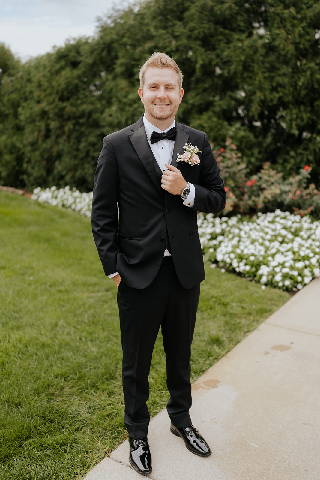 The groom smiles in a black tuxedo and pink rose boutonniere on a garden path lined with white flowers