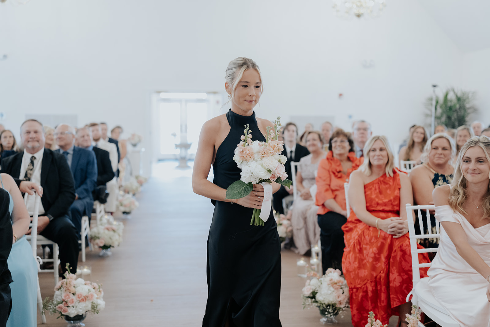 A bridesmaid in a black dress carries a blush bouquet down the aisle as guests watch