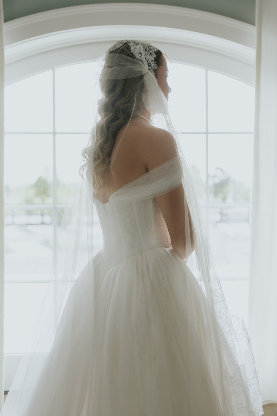 The bride gazes out a tall arched window in her off-the-shoulder gown and lace-trimmed veil.