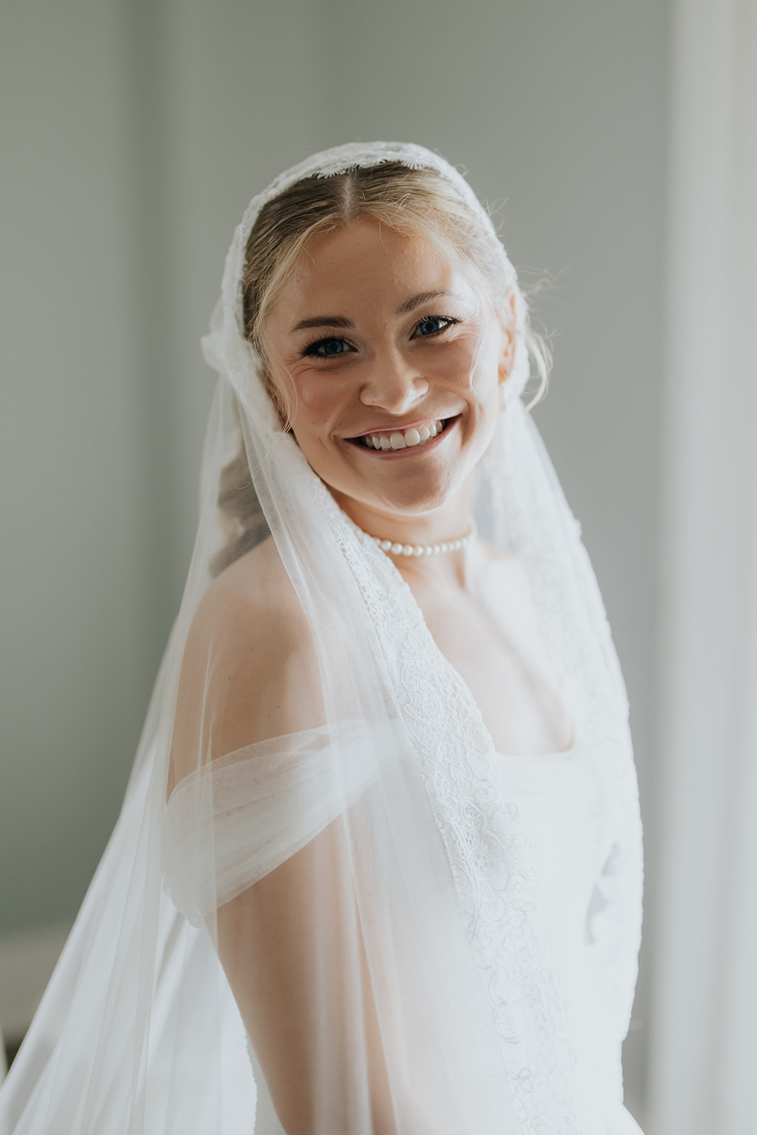 The bride smiles warmly beneath her lace-edged veil, wearing a pearl necklace before the ceremony.
