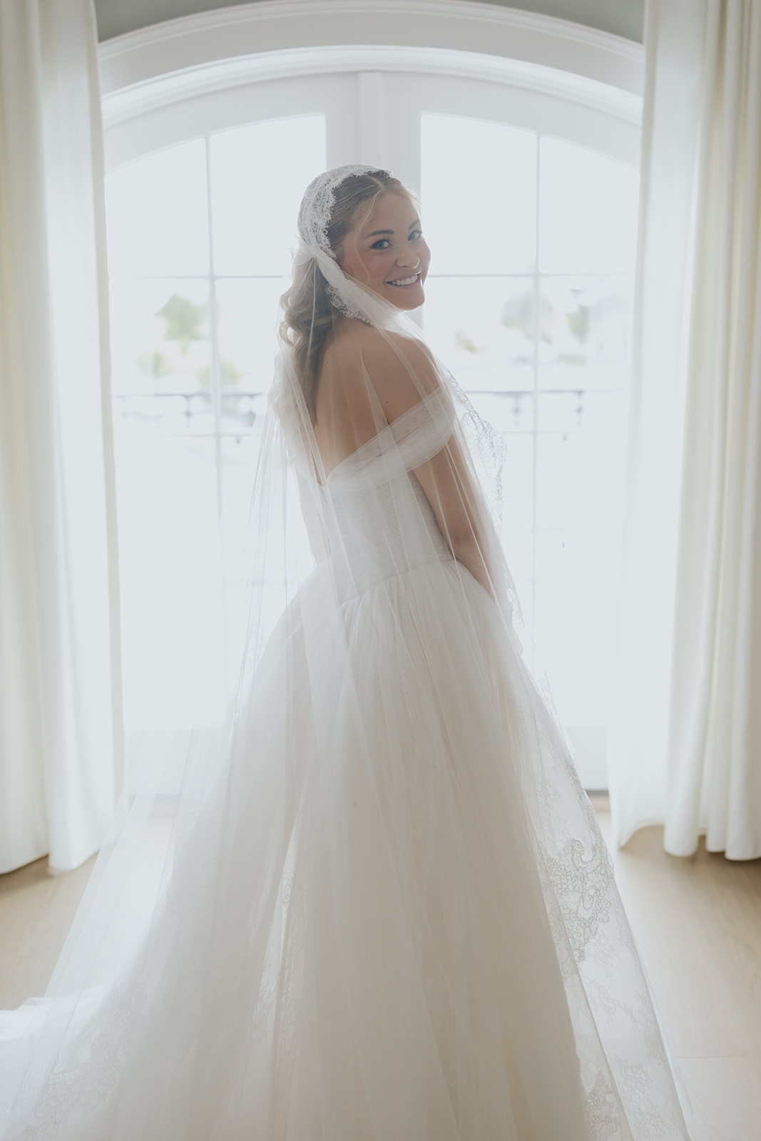 The bride glances back over her shoulder in front of arched windows, her veil trailing behind her.