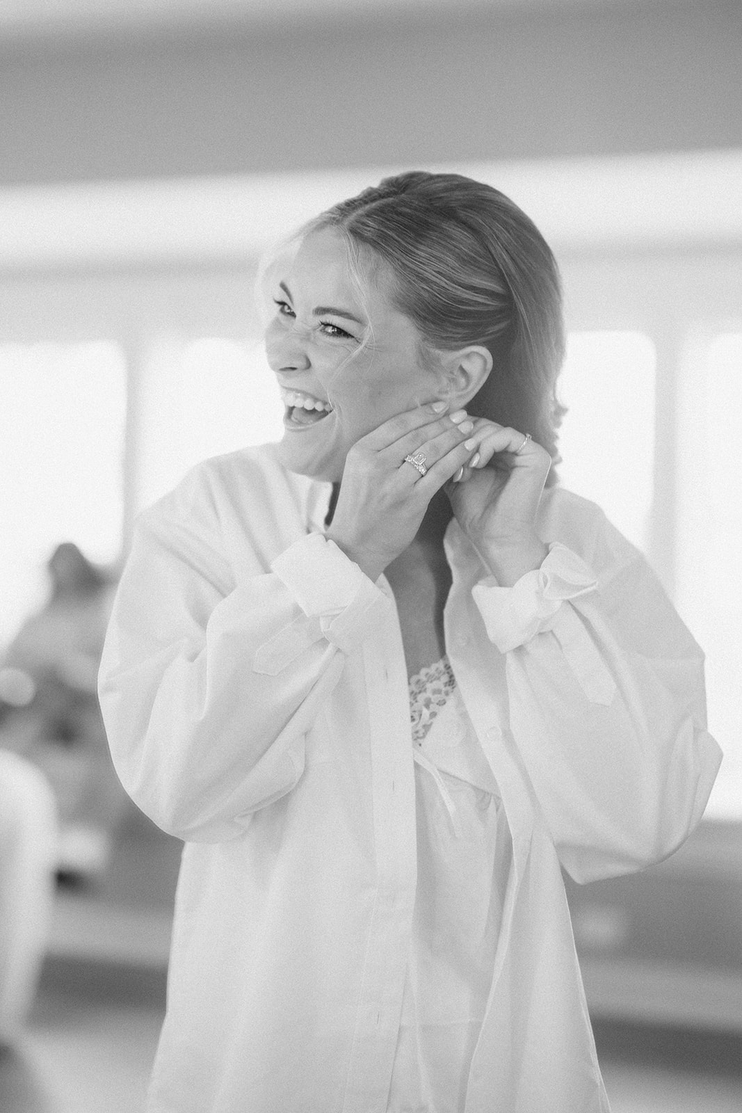 Black and white photo of the bride laughing as she fastens her earrings in an oversized white shirt.