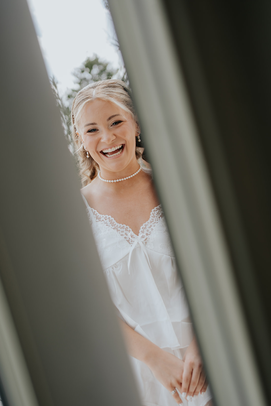 The bride laughs through a partly open door while getting ready in a lace-trimmed white top.
