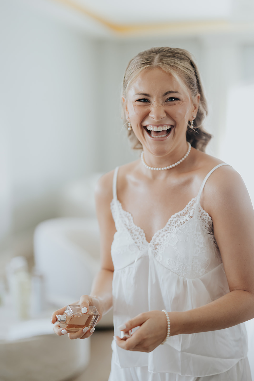 The bride laughs while holding a bottle of perfume as she gets ready in her pearl necklace.