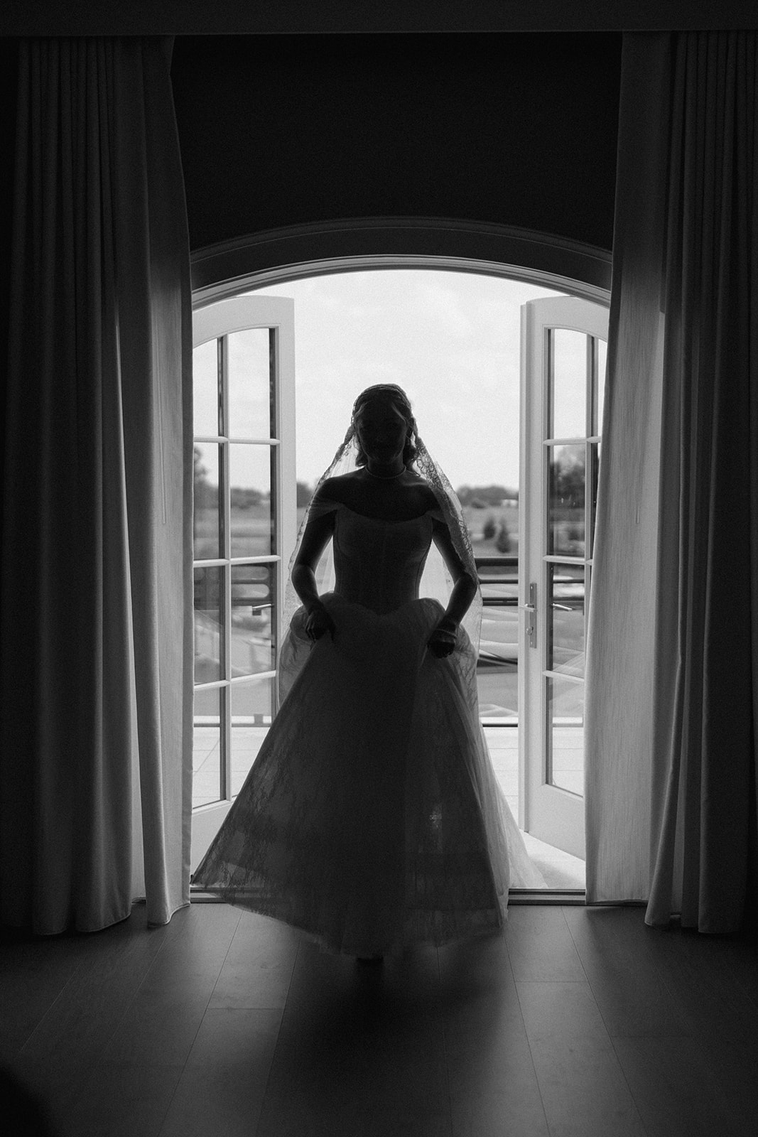 Black and white silhouette of the bride stepping through open arched balcony doors in her gown.