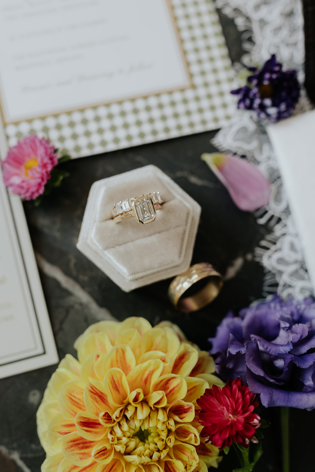Emerald-cut engagement ring in a velvet box beside a gold band, dahlias and the invitation suite.