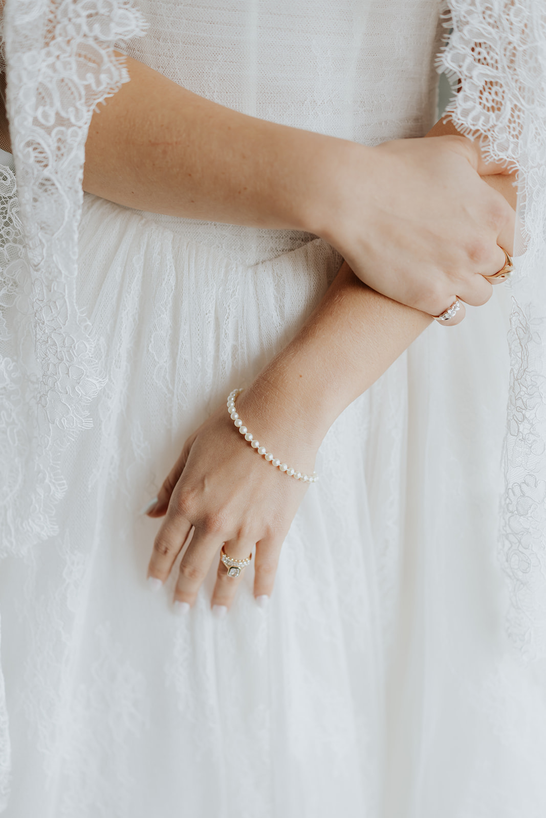 Close-up of the bride's hands with a pearl bracelet and engagement ring against her lace gown.