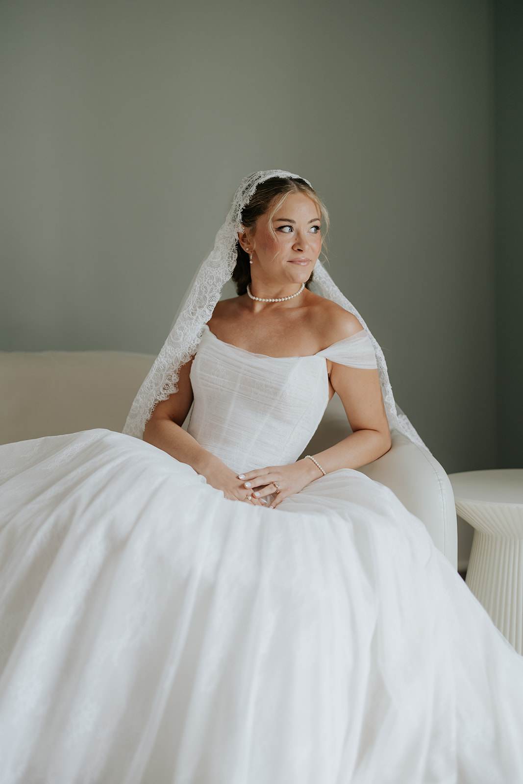 The bride sits on a cream sofa in her full gown and lace veil, gazing toward the window.