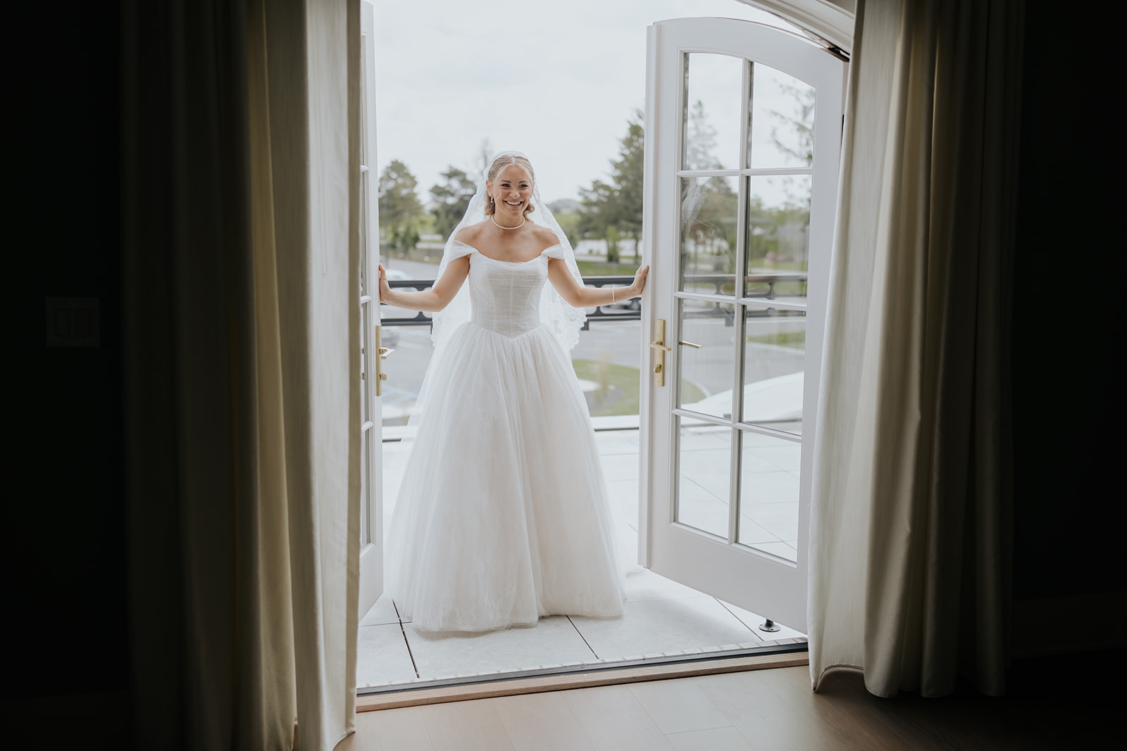 The bride smiles on the balcony as she holds open the white French doors in her ball gown.