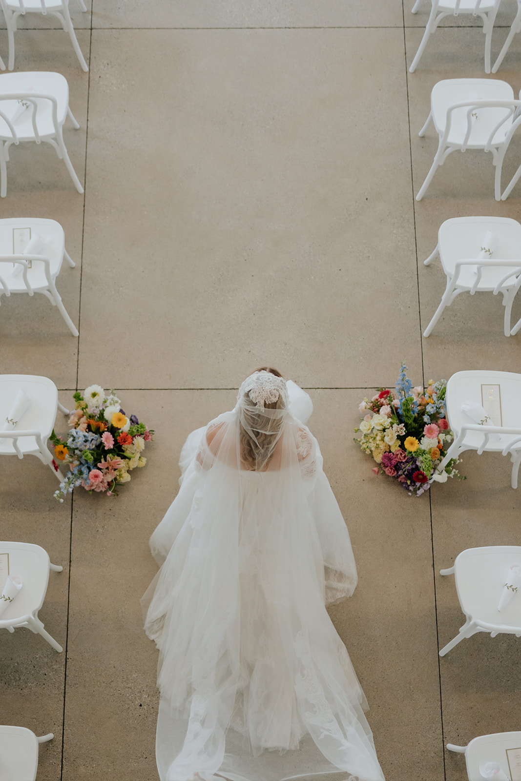 Overhead view of the bride walking between rows of white chairs and colorful floral arrangements.
