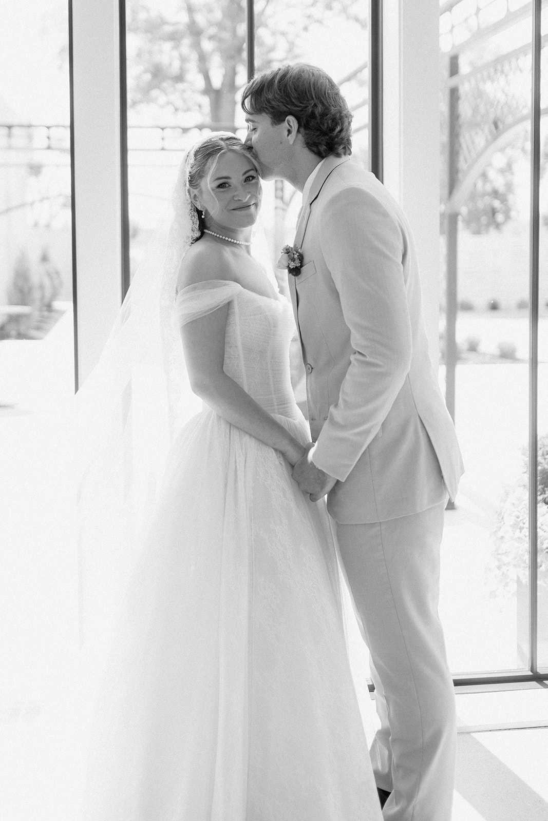 Black and white portrait of the groom kissing the bride's forehead as she smiles at the camera.
