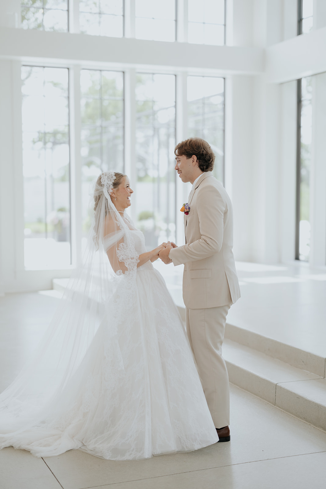 Cami and Trey hold hands and laugh together in a sunlit room lined with tall windows.