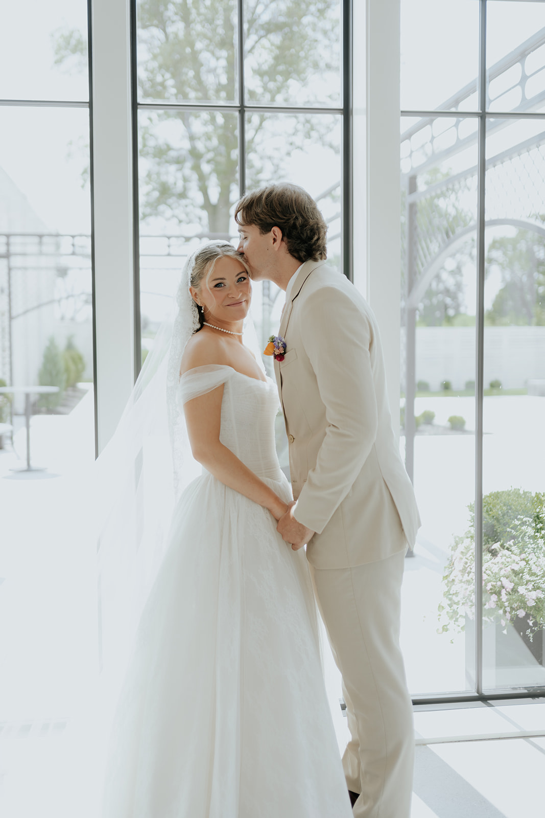 The groom kisses the bride's temple as they stand in front of tall black-framed windows.