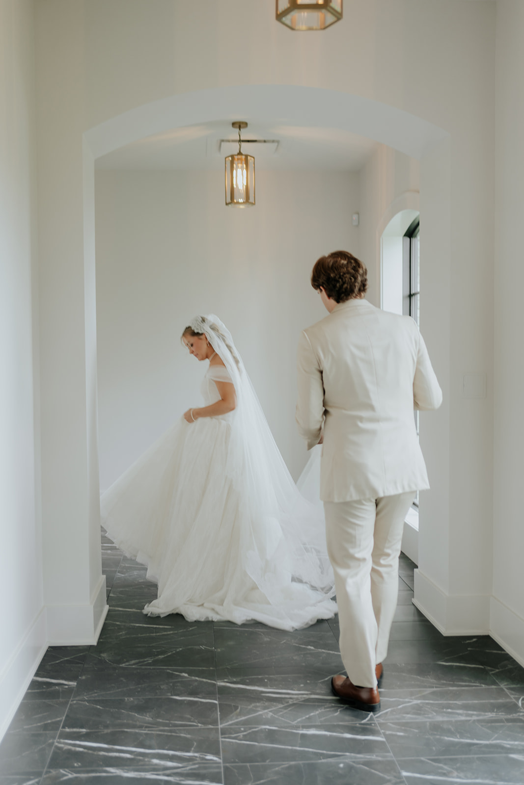 The groom walks up behind the bride in a white arched hallway just before their first look.