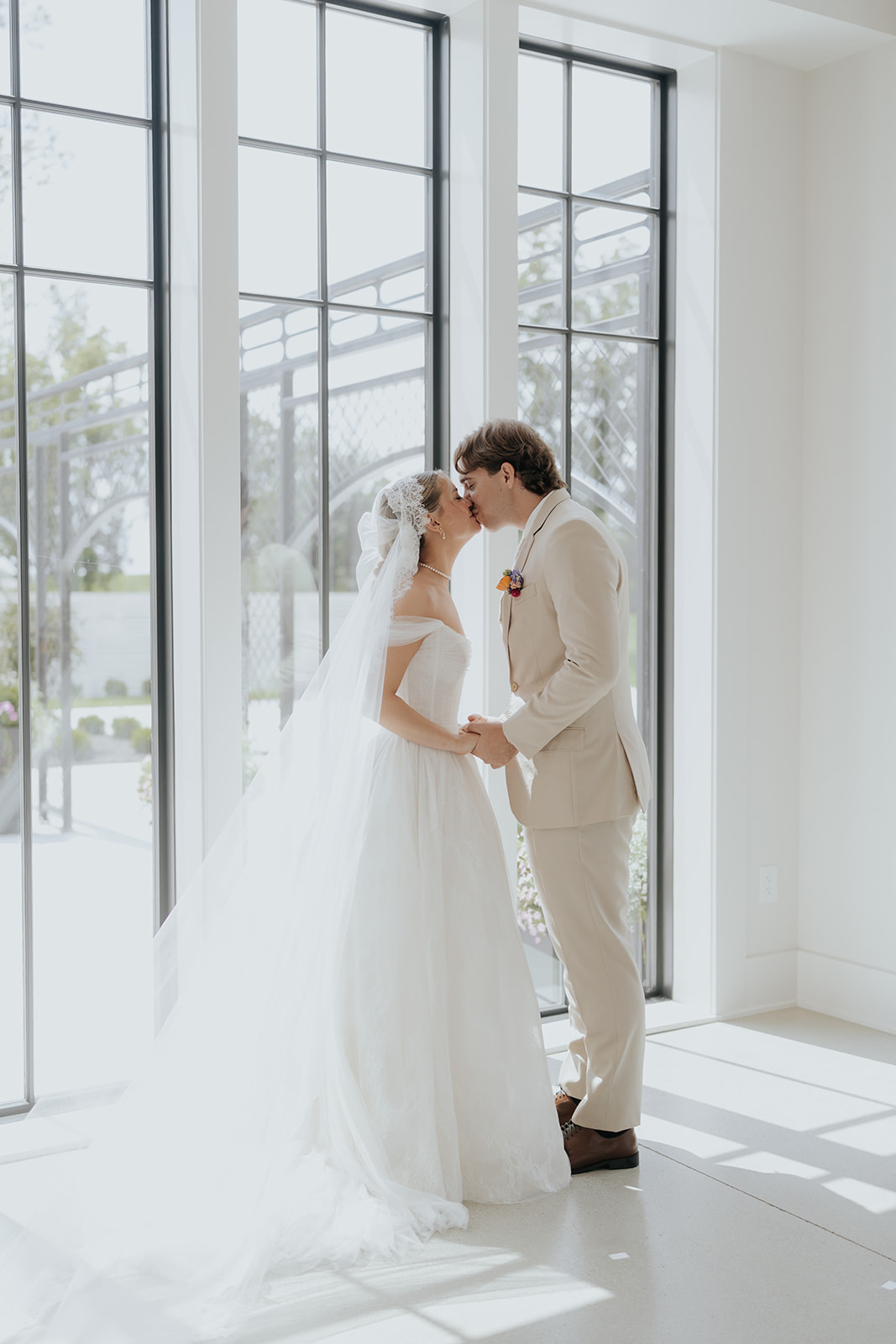 Cami and Trey share a kiss beside tall black-framed windows in soft afternoon light.