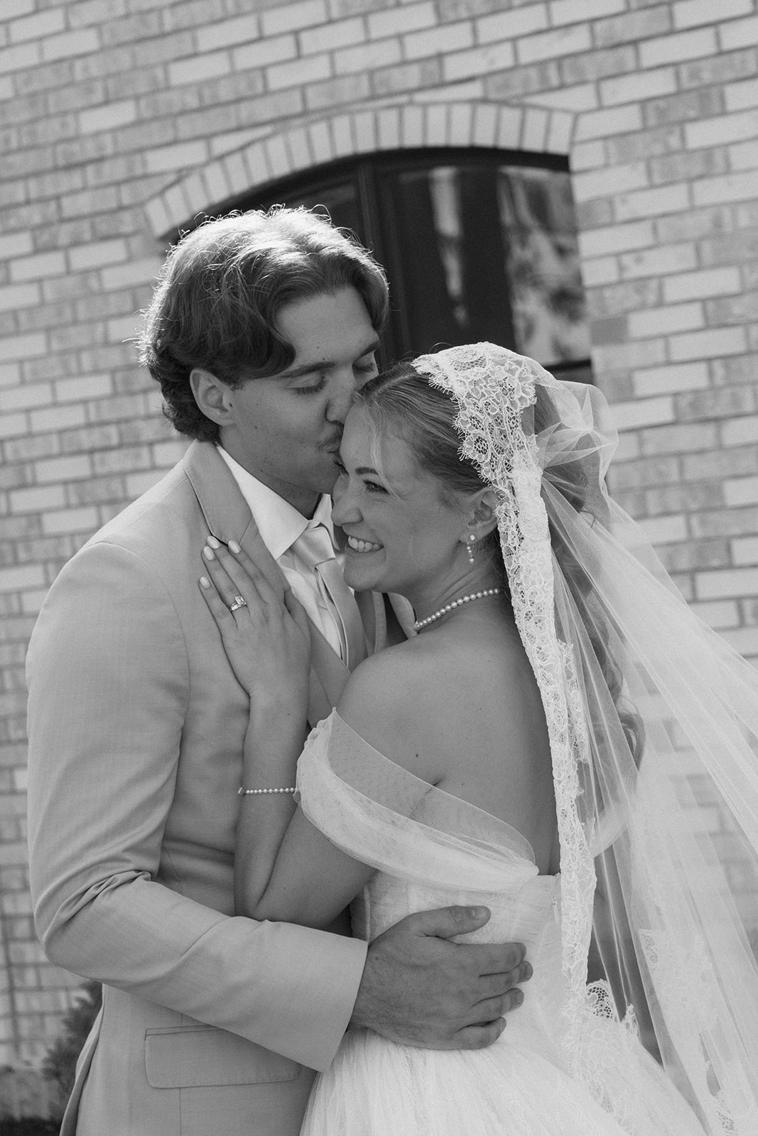 Black and white photo of the groom kissing the bride's cheek as she laughs in her lace veil.