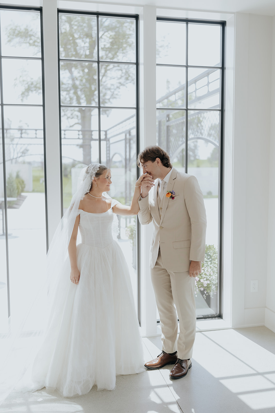 The groom kisses the bride's hand as they stand in front of tall black-framed windows.