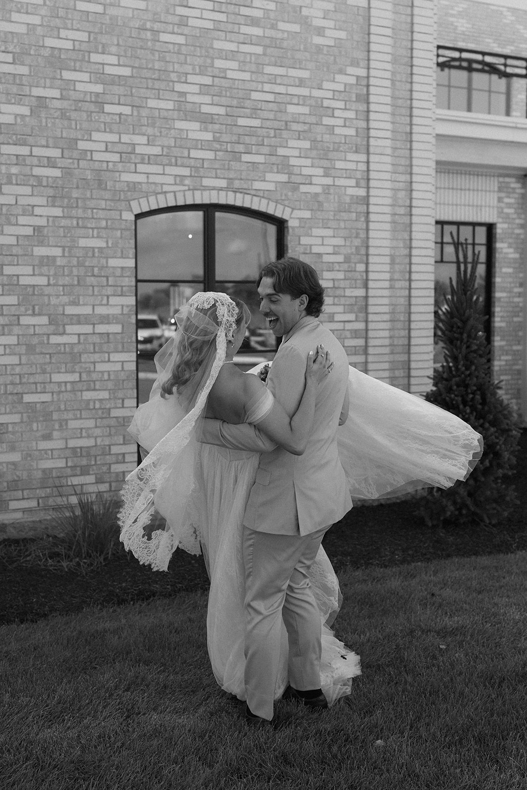 Black and white photo of the groom spinning the bride as her lace veil flies outside a brick building.