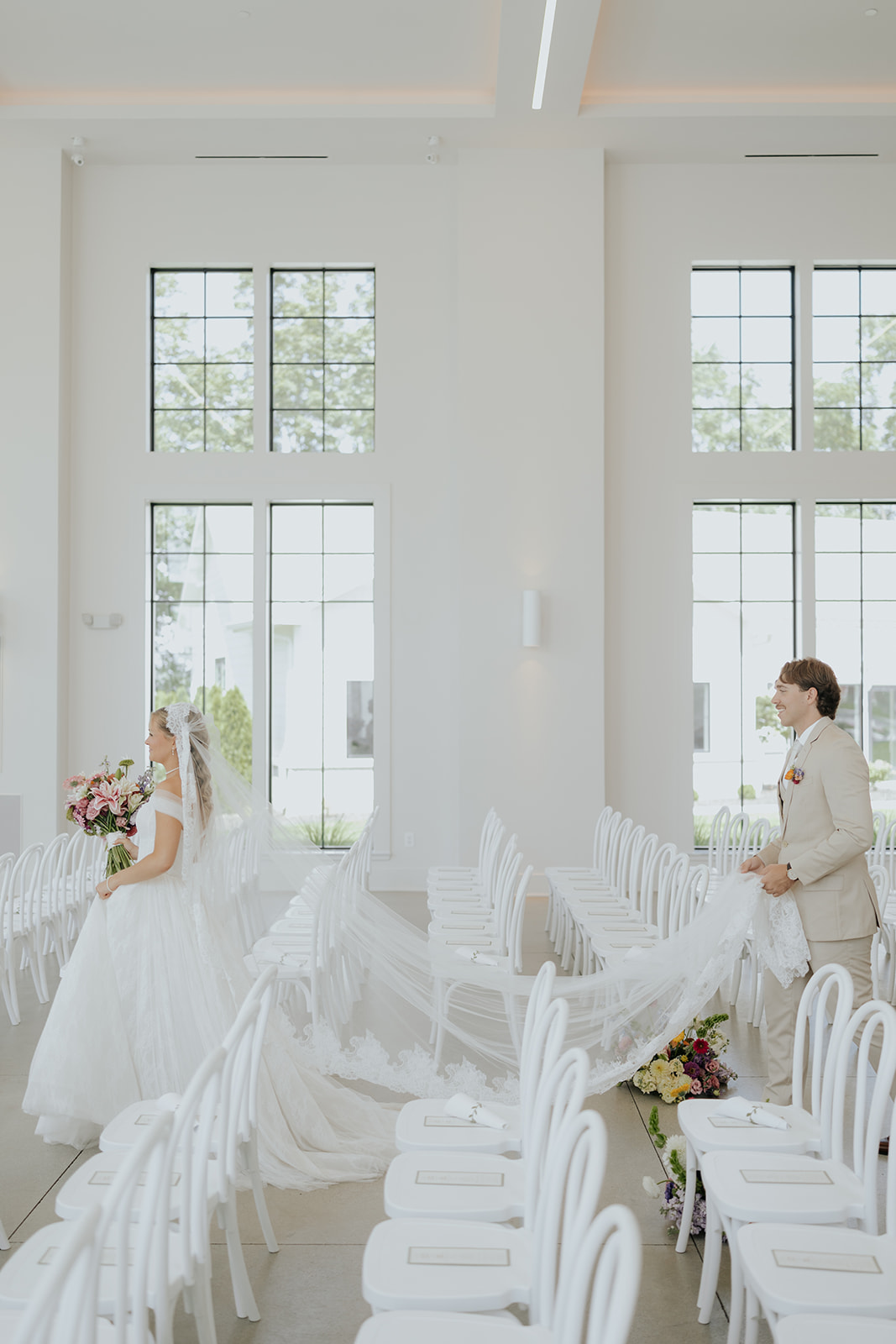 The groom holds the bride's long cathedral veil across the rows of white ceremony chairs.