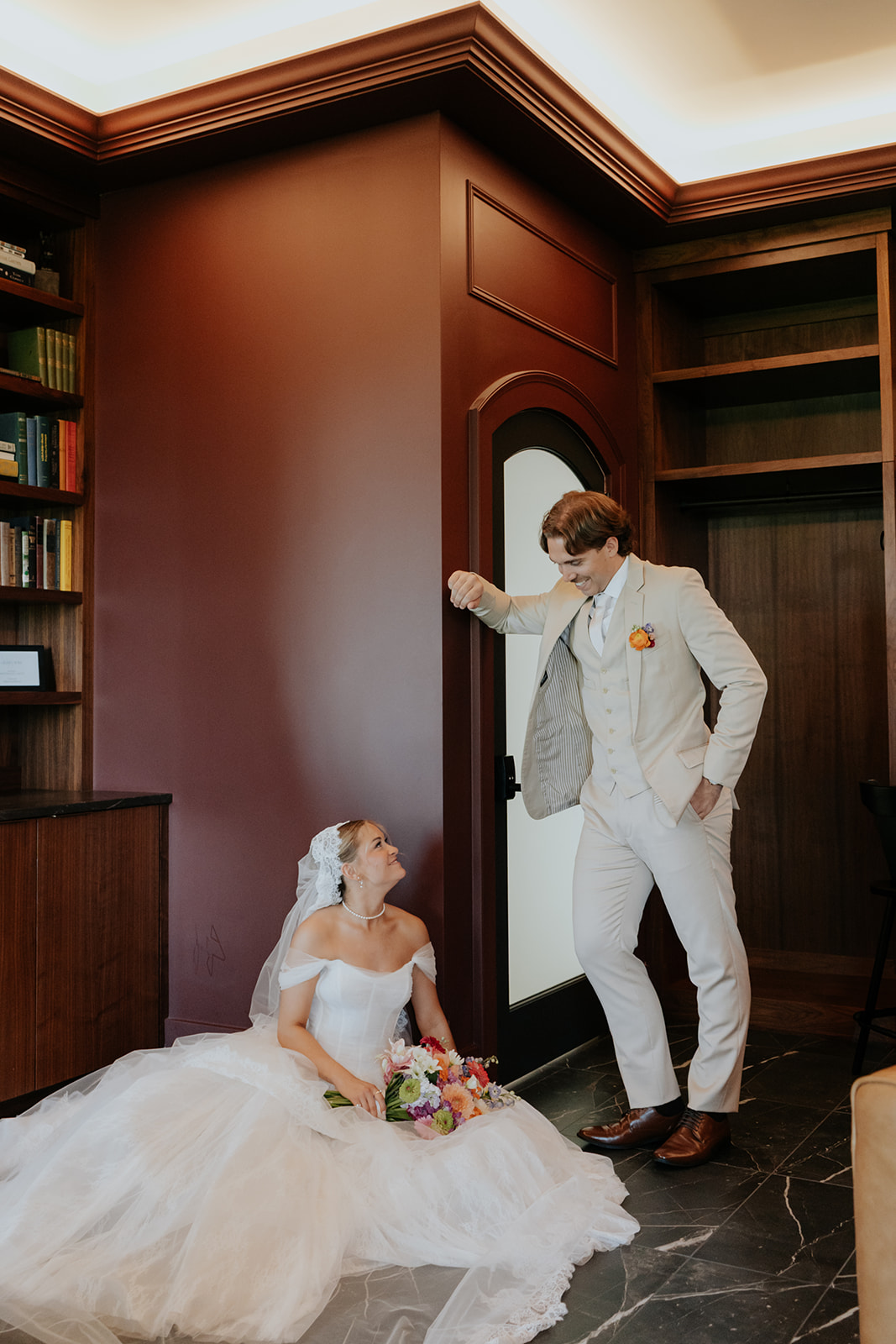 The bride sits on the floor with her bouquet, smiling up at the groom in a wood-paneled library.