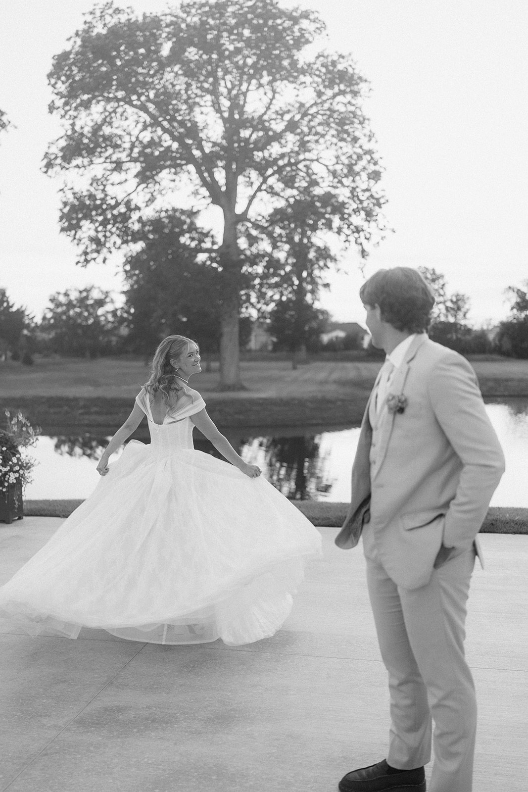 Black and white photo of the bride twirling her gown beside the pond as the groom watches.