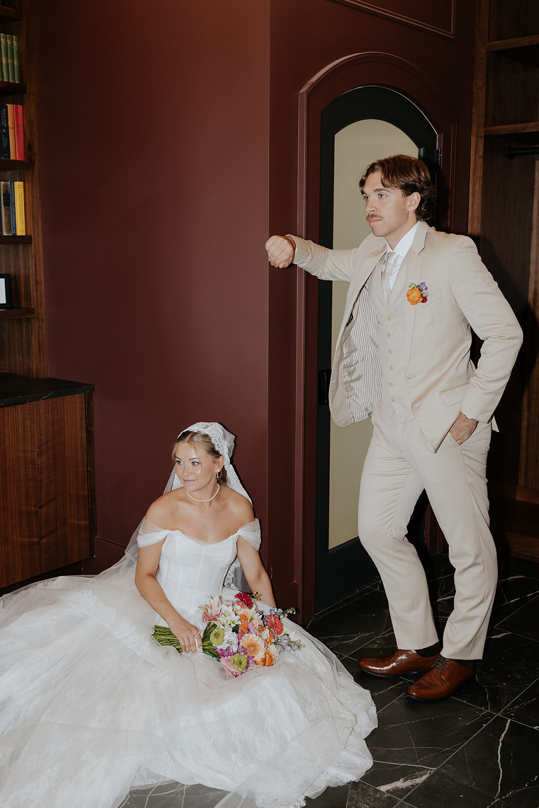 The groom strikes a playful pose by the library door while the bride sits with her colorful bouquet.