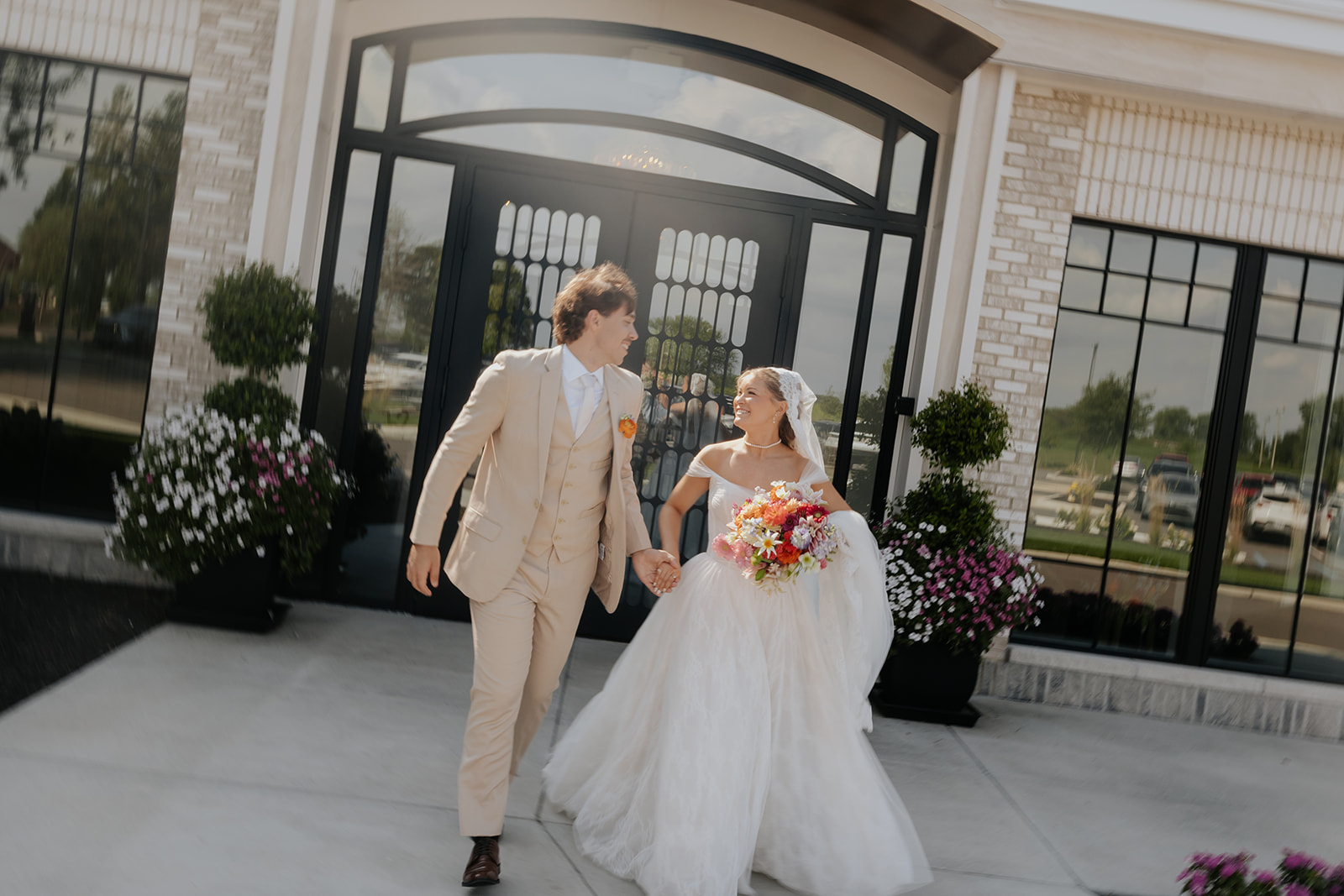 Cami and Trey walk hand in hand out of the arched glass entrance, her bouquet bright with summer color.