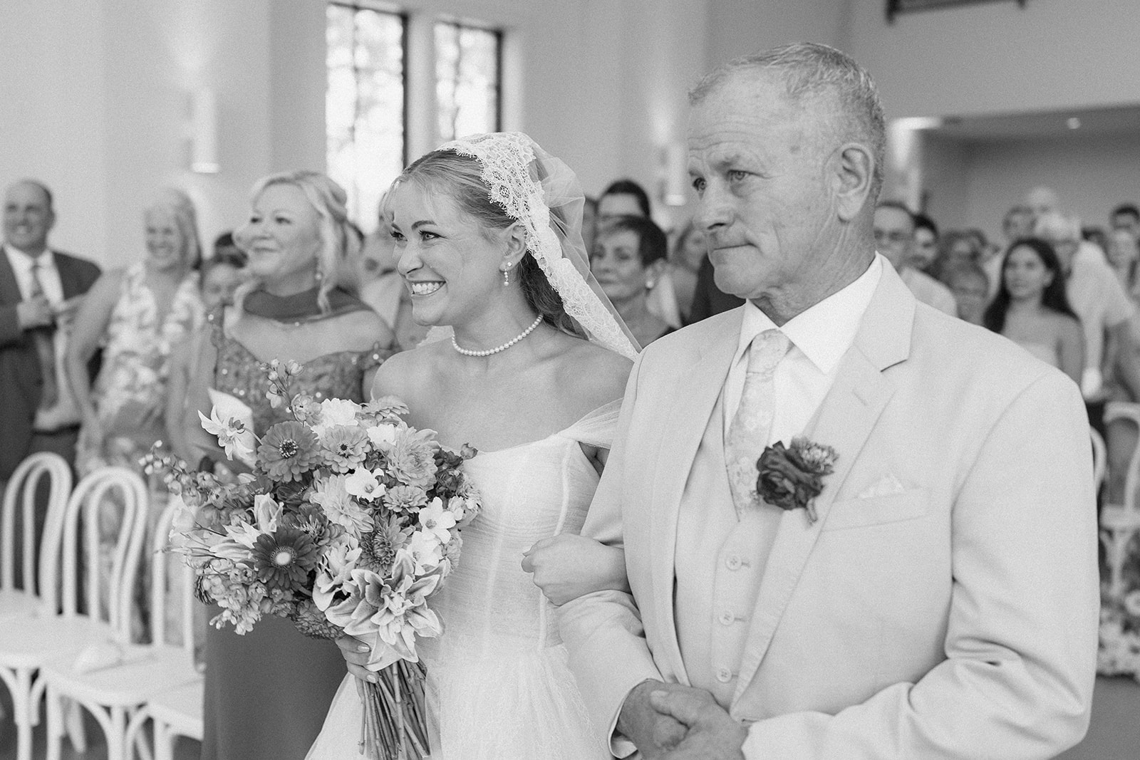 Black and white photo of the bride beaming as she is escorted down the aisle past smiling guests.