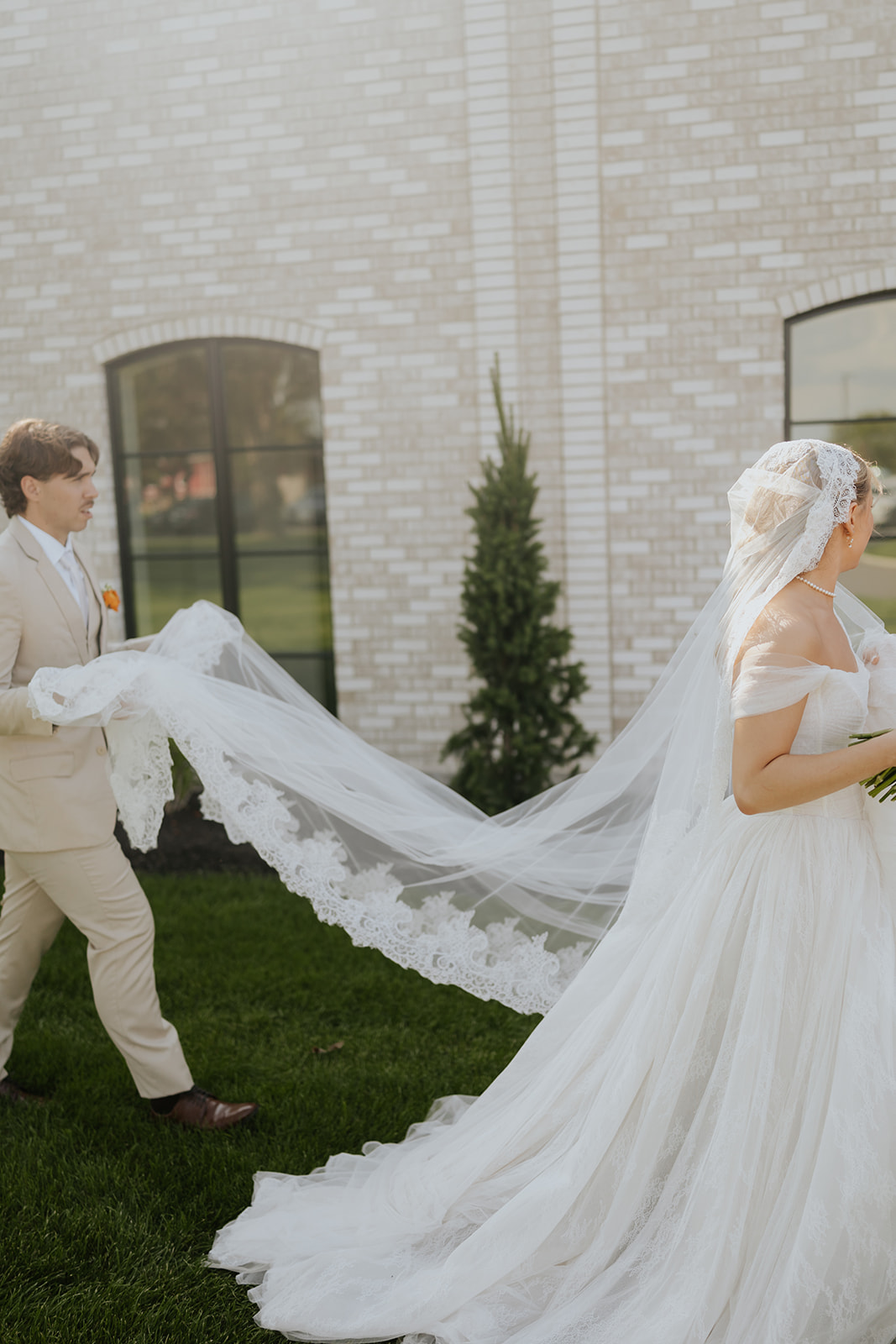 The groom carries the bride's long lace-edged veil as they walk across the lawn.