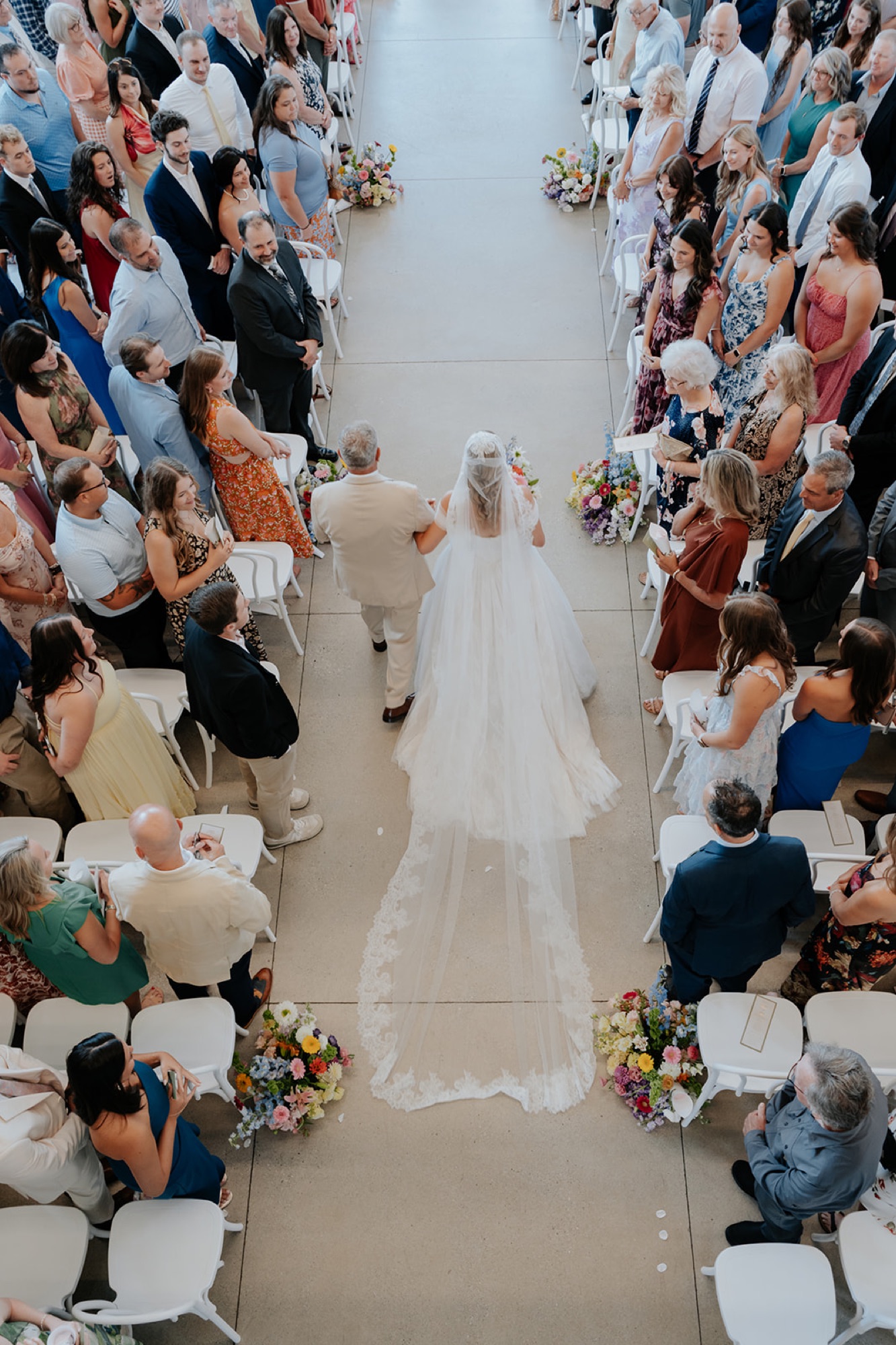 Overhead view of the bride being escorted down the aisle, her cathedral veil trailing past guests.