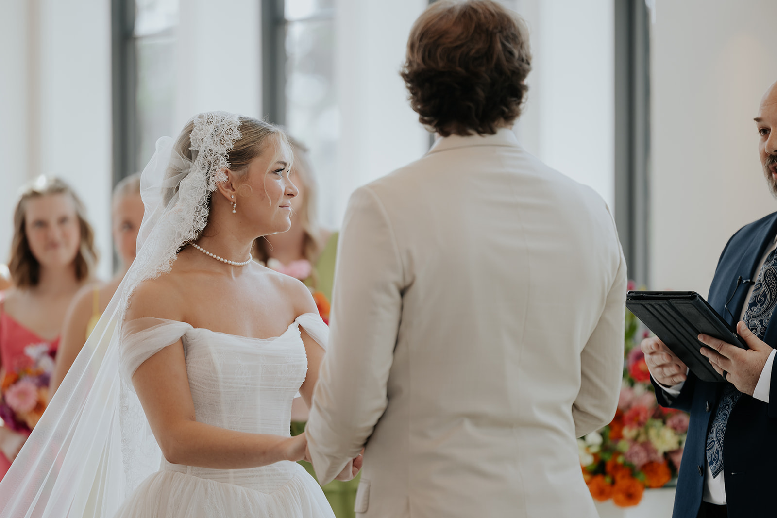 The bride smiles at the groom as they hold hands during the vows, the officiant reading beside them.