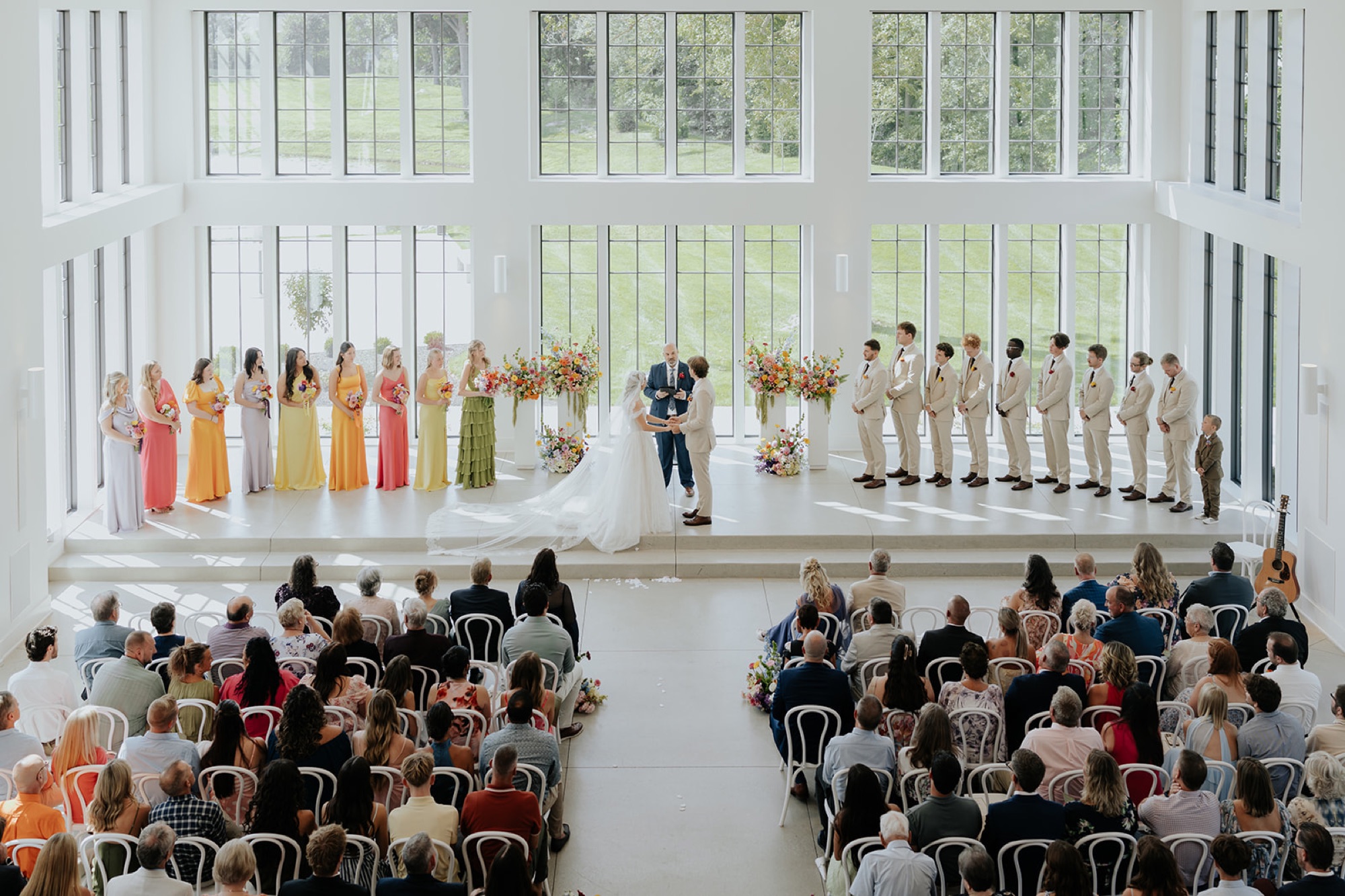 Wide view of the ceremony beneath towering windows, with bridesmaids in bright colors and groomsmen in tan suits.