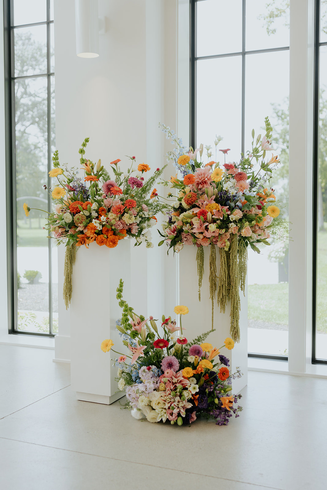 Tall ceremony arrangements of orange, pink and yellow flowers stand on white pedestals by the windows.