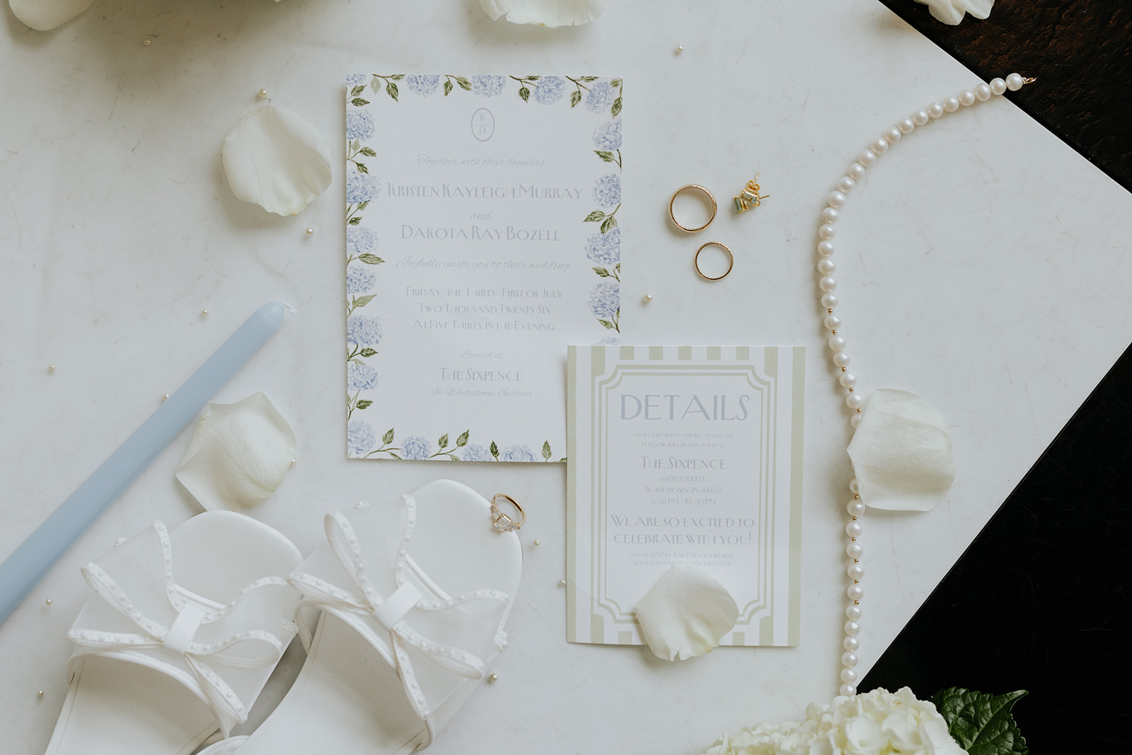 Blue hydrangea wedding invitation, details card, rings, pearls and bridal shoes laid flat on white marble