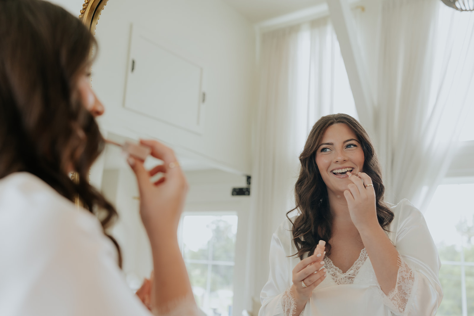 The bride smiles at her reflection in a gold mirror as she touches up her lip gloss in a lace robe