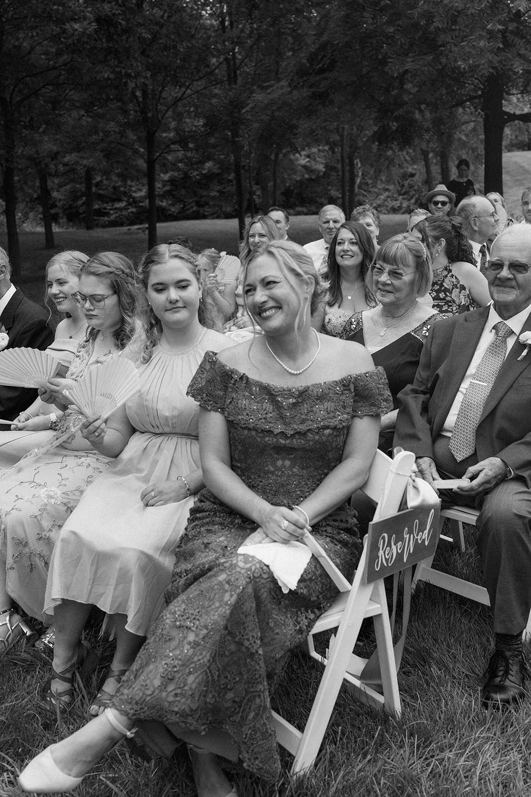 Black and white photo of smiling guests seated in white chairs at an outdoor ceremony in the trees