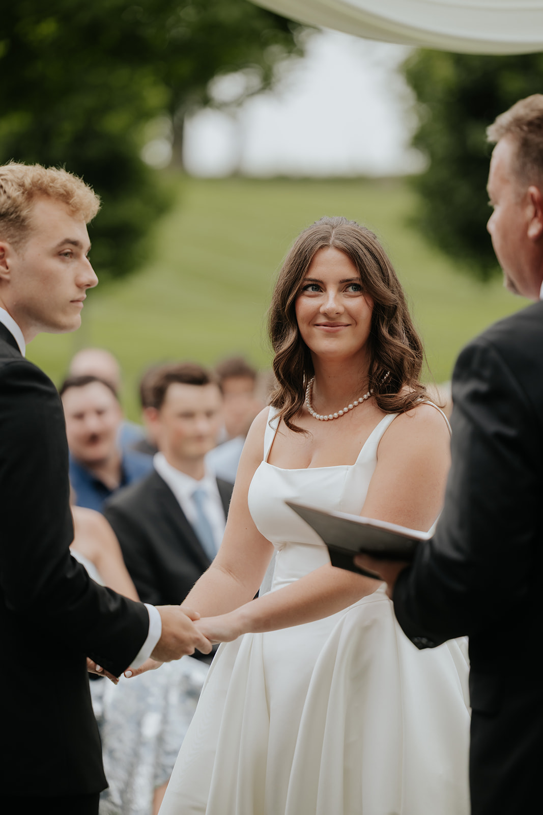 Kristen smiles at Cody as they hold hands during their outdoor vows with guests behind them
