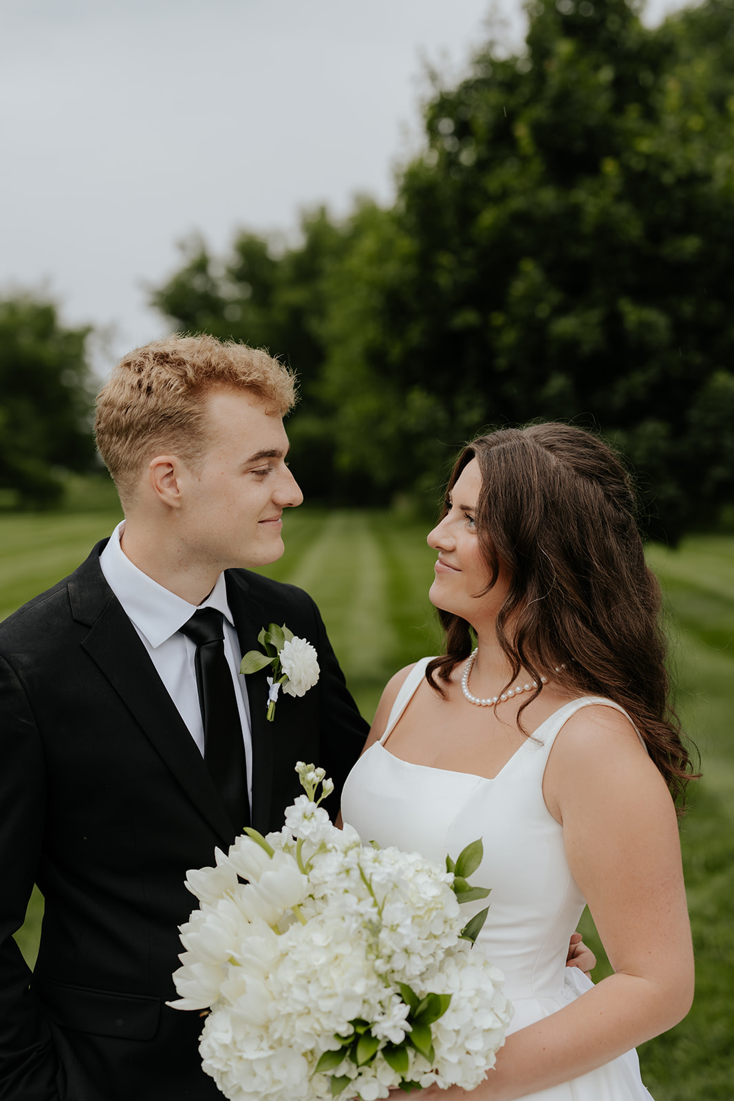 Cody and Kristen gaze at each other holding a white hydrangea bouquet on a striped green lawn