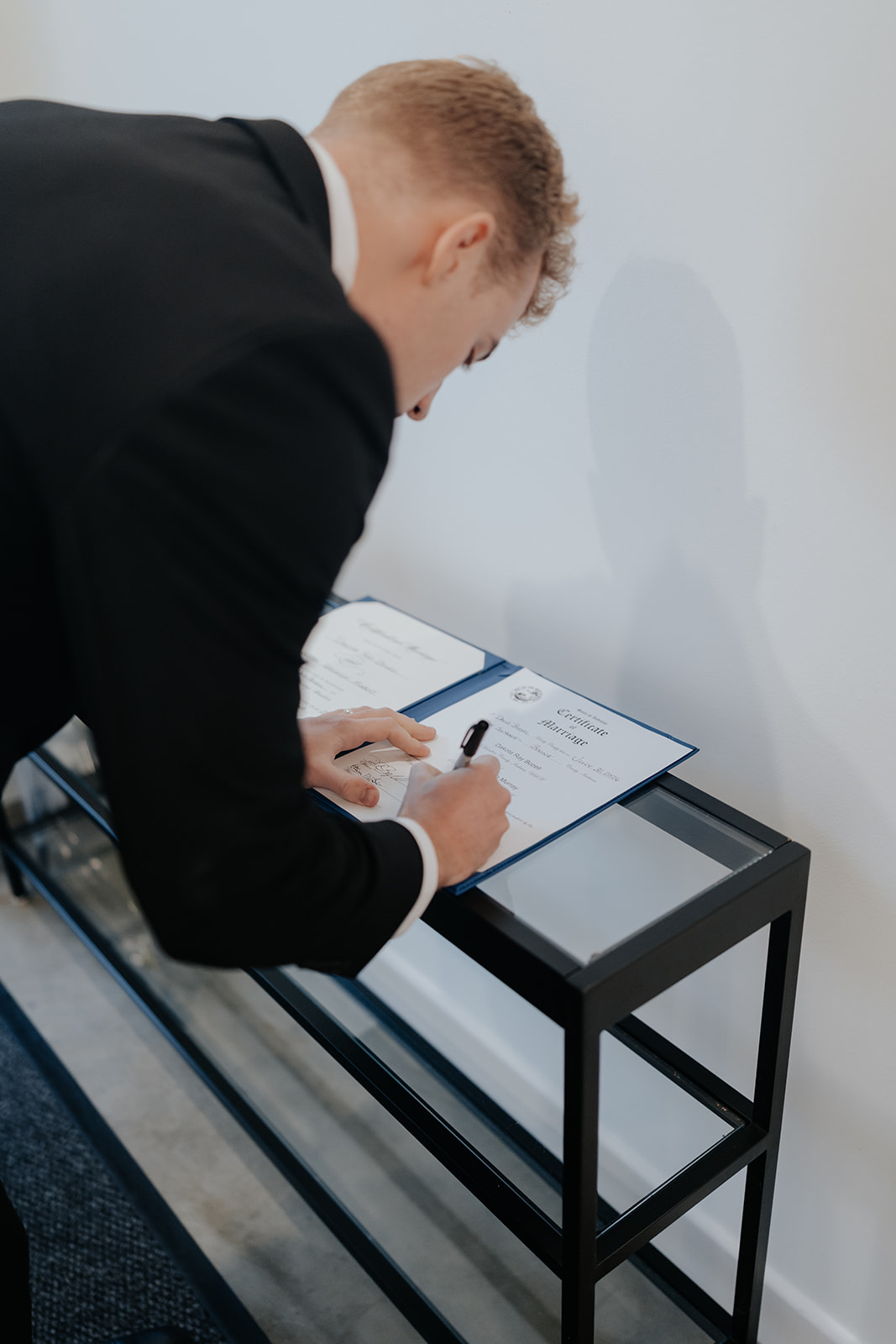 The groom leans over a black side table to sign the marriage certificate