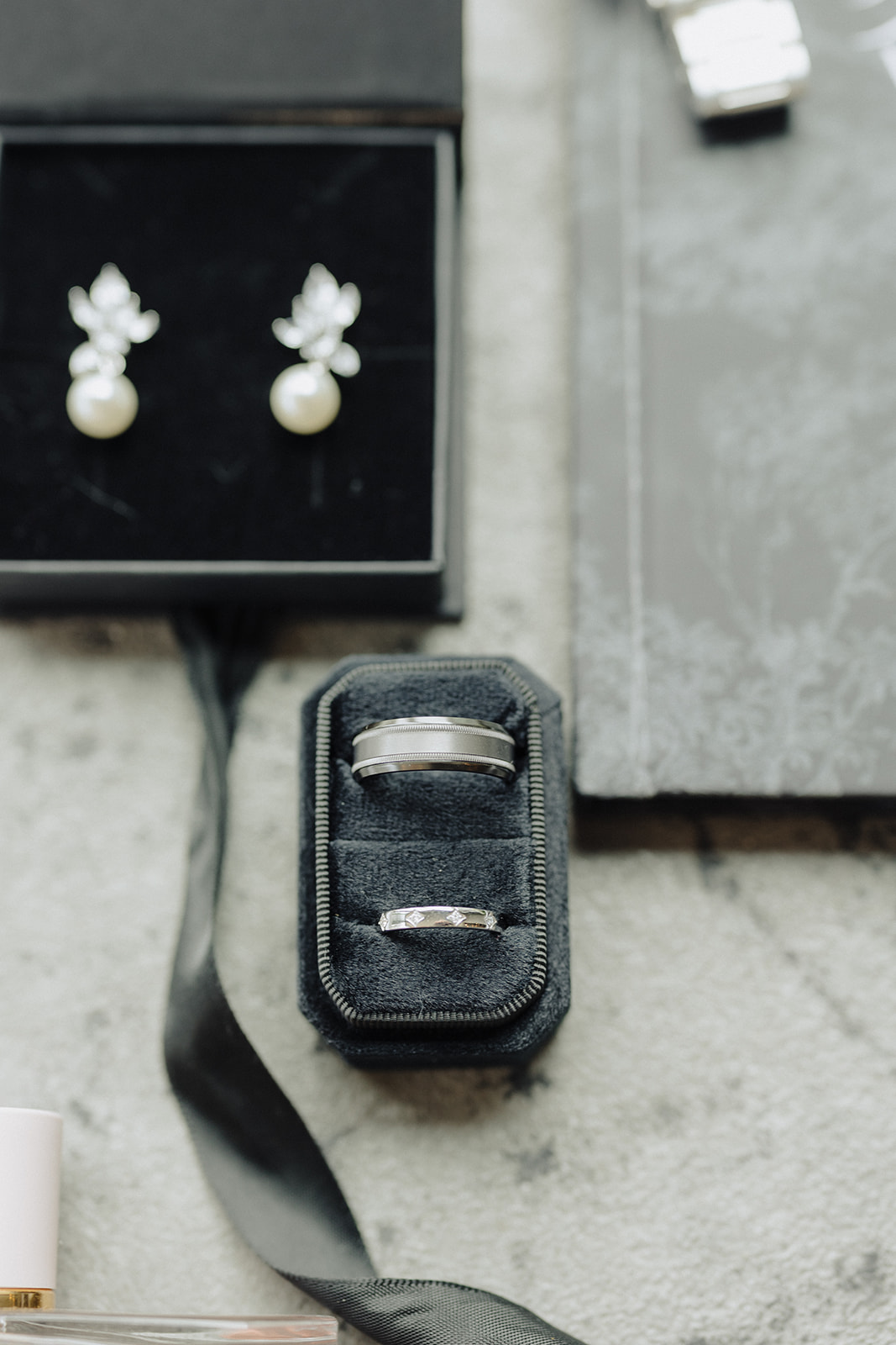 Pearl drop earrings in a black box beside wedding bands in a velvet ring box