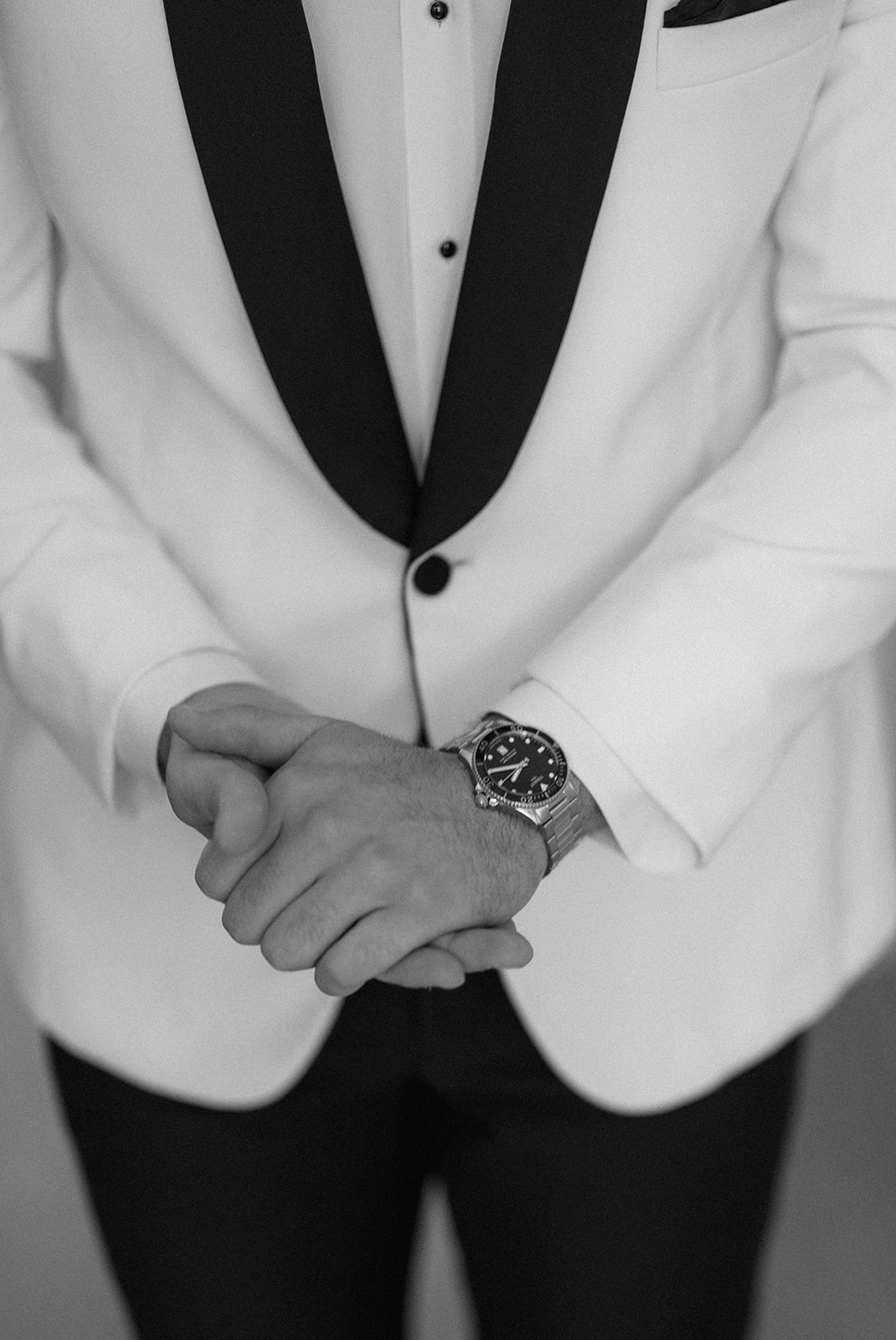 Black and white close-up of the groom's clasped hands and watch against his white dinner jacket