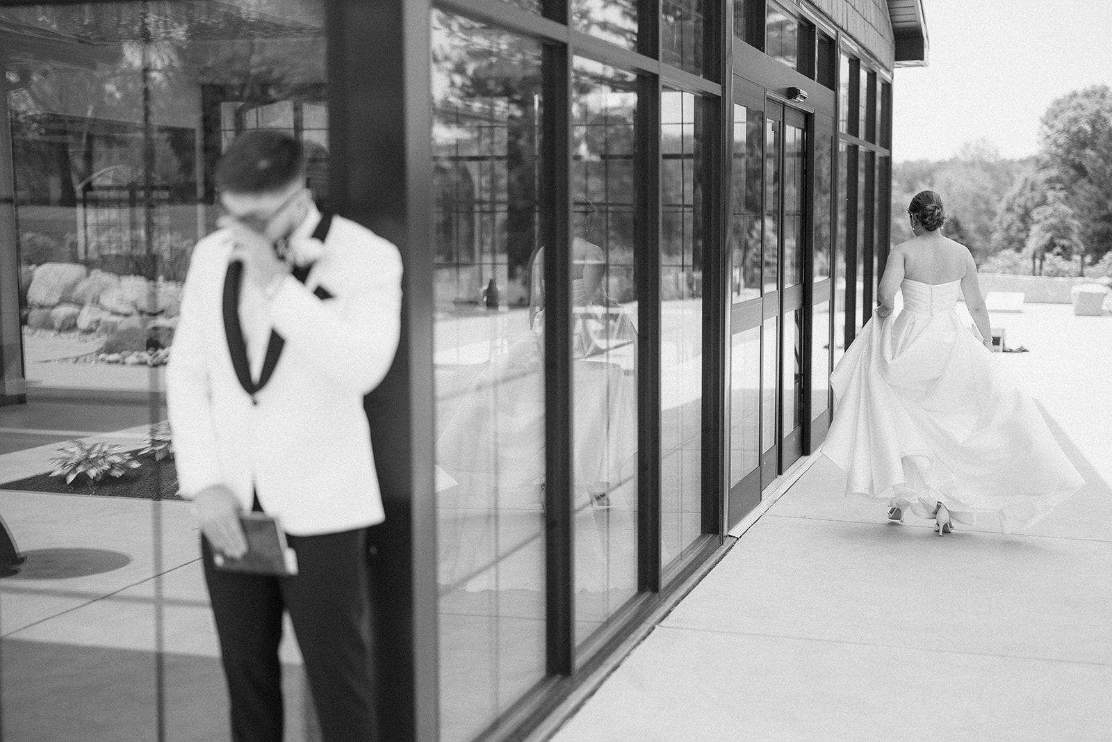 Black and white photo of the groom wiping a tear as the bride walks away along the glass wall