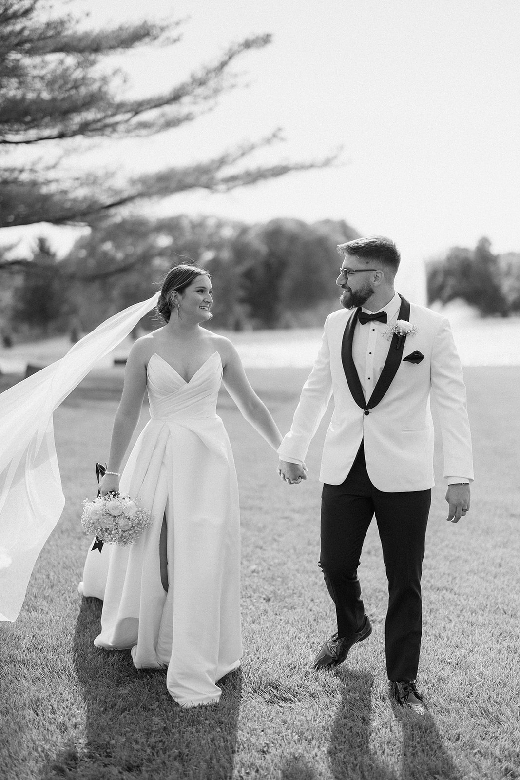 Black and white photo of Dallas and Beau walking hand in hand as her veil lifts in the wind