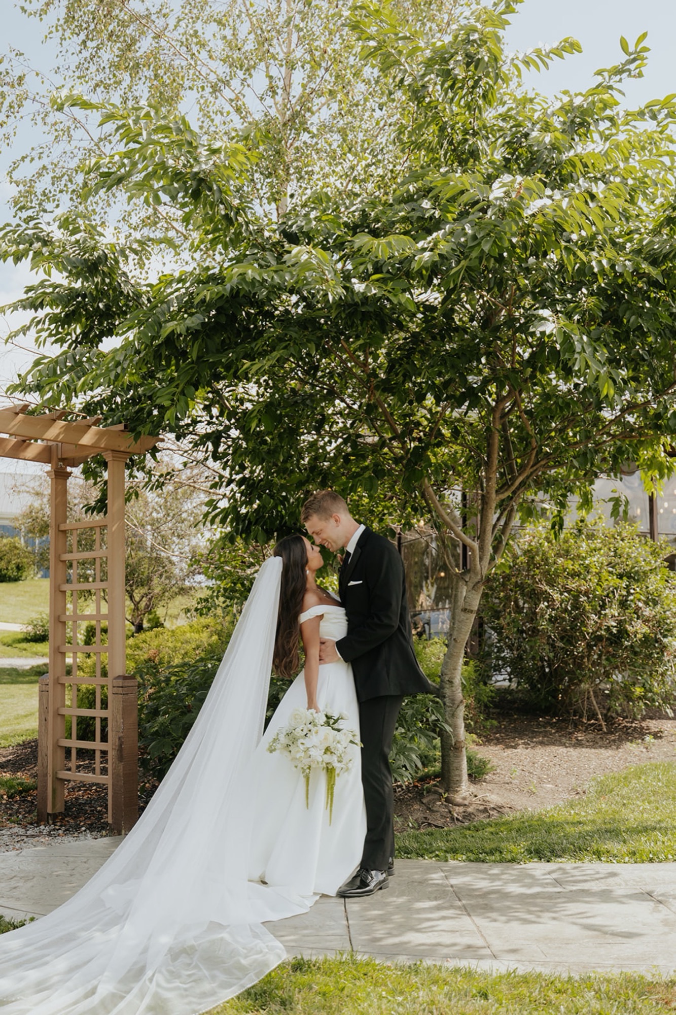 Isabel and Benjamin share a kiss beneath a leafy tree beside a wooden garden arbor.